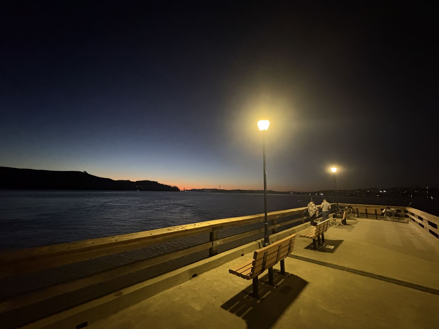 The Benicia fishing pier after sunset, lamps lit along the railing and anglers at the far end, with the Carquinez Strait and the hills beyond.